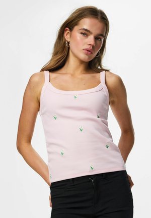 Jeune femme portant un débardeur côtelé rose avec une petite broderie de fleurs blanches et un pantalon noir, posant sur un fond blanc uni.