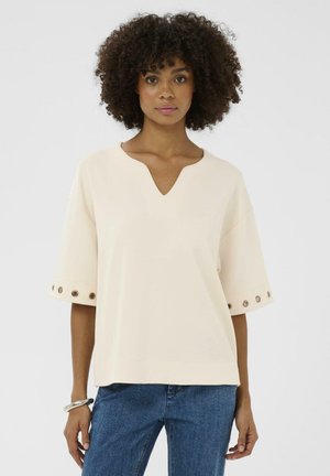 Kremowy, krótki top z krótkim rękawem, dekoltem w serek i ozdobnymi metalowymi nitemi przy mankietach. Gładka faktura, luźny krój i casualowy design.