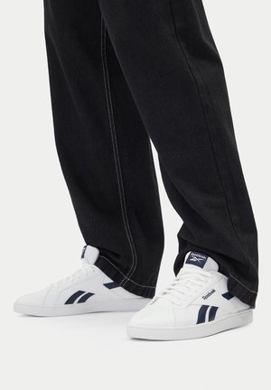 Białe sneakersy Reebok z granatowym logo, noszone z czarnymi spodniami z widocznym białym przeszyciem na jednolitym białym tle.