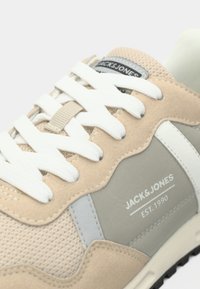 Μπεζ αθλητικό παπούτσι με γκρι λεπτομέρειες, υλικά από δίχτυ και σουέτ, άσπρα κορδόνια και λογότυπα. Διαθέτει υφή στη σόλα και στιβαρό, πολυεπίπεδο σχέδιο.