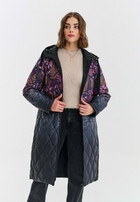 Žena v dlouhém prošívaném kabátu s fialovými květinovými vzory, béžovém svetru, černých džínách a se zlatým přívěskem se dívá na stranu.