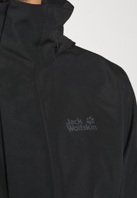 Veste noire imperméable avec col zippé, arborant un logo discret sur la poitrine. Le matériau semble lisse avec une finition mate.