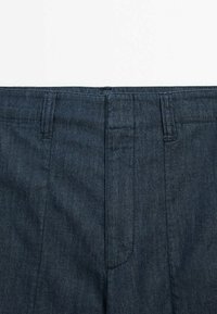Dunkelblaue Denim-Hosen verfügen über eine glatte Textur, ein Fünf-Taschen-Design und kontrastierende Nähte. Der Bund enthält Gürtelschlaufen zur Anpassung.