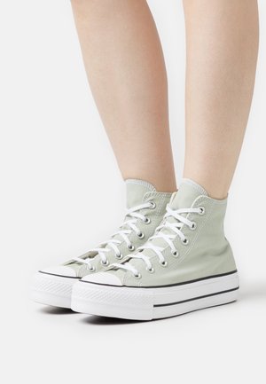 Piernas vistiendo zapatillas de lona de caña alta de color verde claro con cordones blancos y suelas de goma blanca gruesa sobre un fondo blanco.
