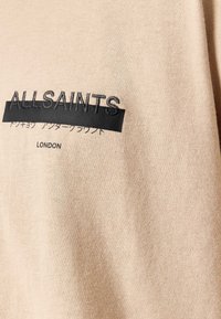 Beżowa tkanina z czarną haftem i nadrukiem zawierającym słowo ALLSAINTS, japońskie znaki oraz tekst LONDON.