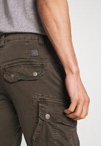 Cargo pants in donker olijfgroen canvas, met opgestikte zakken met drukknoopsluitingen en een stoffen label op de achterkant. Glad textuur.