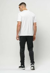 Mann iført hvit t-skjorte og svarte treningsbukser med joggesko, stående og vendt bort fra kameraet mot en ensfarget lys bakgrunn.