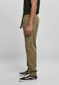 Pantalones de algodón color caqui con cintura elástica y cordón negro. Combinados con zapatillas negras y blancas; mostrados desde un ángulo lateral.
