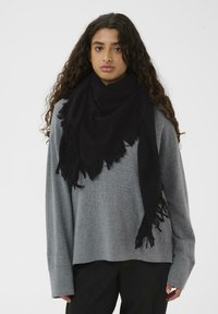 Sort frynset tørklæde lavet af blødt stof, hængende løst om halsen. Båret over en grå, oversized sweater.