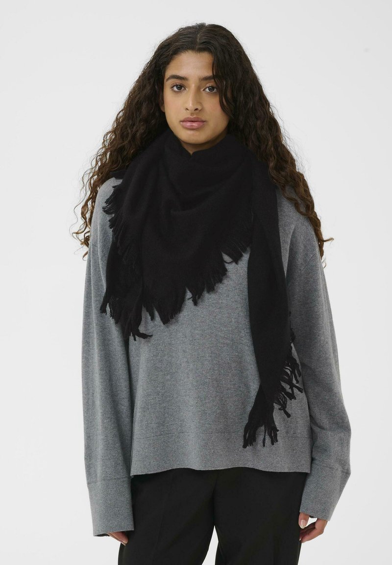 Sort frynset tørklæde lavet af blødt stof, hængende løst om halsen. Båret over en grå, oversized sweater.