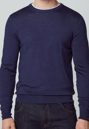 Homme portant un pull ras du cou bleu marine ajusté sur un sous-vêtement blanc, associé à un jean bleu foncé.