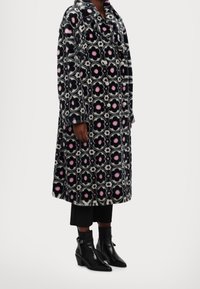 Cappotto in pelliccia sintetica con stampa floreale in nero, rosa e bianco. Presenta un design doppiopetto e un ampio colletto. Indossato con stivali neri alla caviglia.