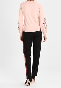 Hellrosa Pullover mit Blumenstickerei auf den Ärmeln, kombiniert mit schwarzen Hosen mit roten Seitenstreifen und schwarzen High Heels.