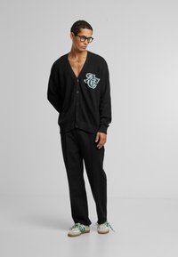 Svart strikket cardigan med V-hals, knappelukking og et kontrastfarget brodert logo. Sammen med svarte bukser og loafers.