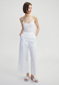 Pantalons en coton blancs avec des découpes brodées de fleurs, design à jambe large, associés à un top en coton blanc et des sandales blanches texturées.