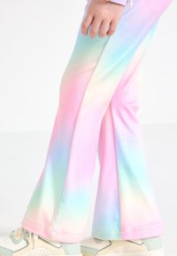Enfant portant un pantalon pattes d'éléphant tie-dye arc-en-ciel pastel et des baskets de couleur claire, debout sur un fond blanc.
