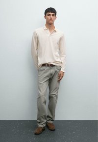 Junger Mann, der gegen eine graue Wand steht und ein cremefarbenes Langarmpolo, graue Jeans, einen braunen Gürtel und braune Wildlederschuhe trägt.