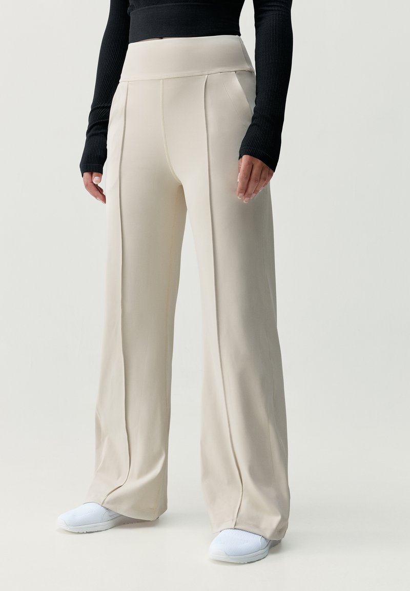 Beige, magas derekú nadrág széles lábakkal, sima anyaggal, függőleges varrással, fehér sportcipővel párosítva.