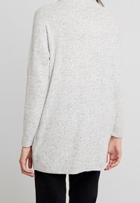 Hellgrauer Pullover aus einem weichen, strukturierten Stoff, mit langen Ärmeln, lockerer Passform und abgerundetem Saum, getragen über dunklen Hosen.
