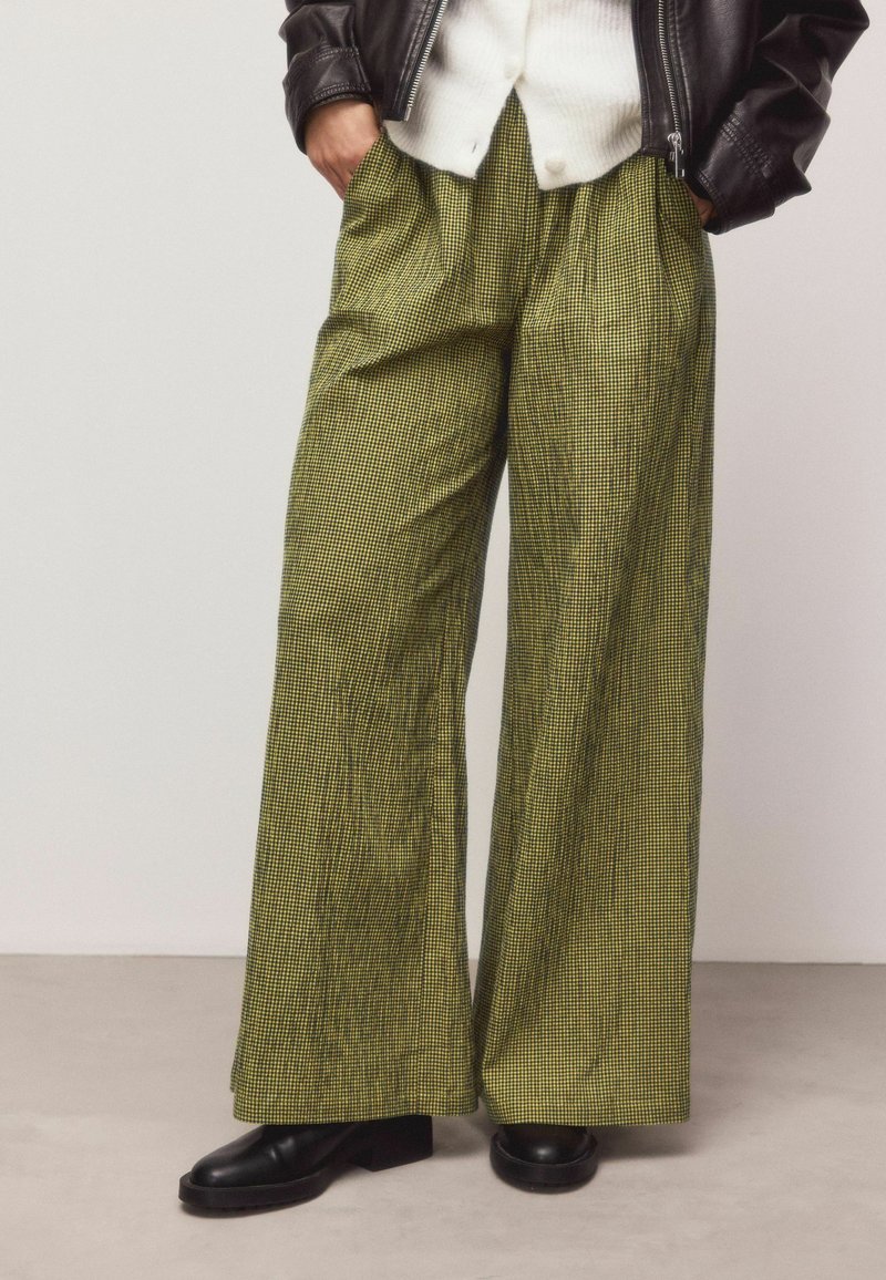 Pantalon à jambes larges à carreaux verts avec une finition texturée et des poches latérales, associé à des chaussures à plateforme noires et une veste en cuir sombre.