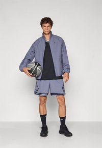 Casaco atlético cinza leve com gola alta, top de alças preto, calções cinza com padrão e ténis de basquetebol pretos, segurando uma bola de basquetebol.