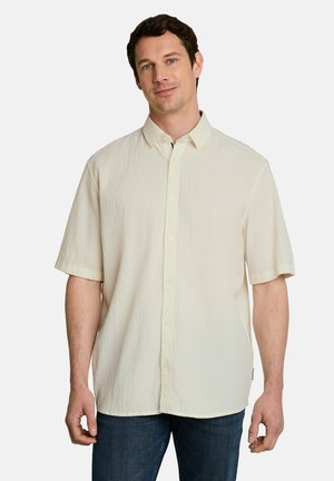 Hombre con una camisa crema de manga corta con botones y vaqueros azul oscuro, de pie frente a un fondo claro y liso.