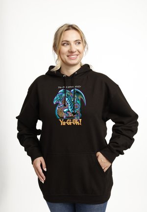 Kvinde smiler og bærer en sort hættetrøje med en blå drage og teksten "Yu-Gi-Oh!" foran mod en hvid baggrund.