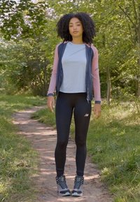 Sportoutfit mit einem hellblauen Oberteil, rosa Ärmelakzenten, schwarzen Leggings und strukturierten schwarzen Sneakers mit grünen Sohlen, auf einem natürlichen Weg.