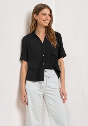 Femme debout souriante, portant une chemise noire à manches courtes boutonnée et un pantalon blanc à cordon devant un mur clair uni.