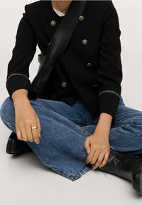 Czarna marynarka dwurzędowa z złotymi guzikami, zestawiona z niebieskimi szerokimi spodniami jeansowymi, noszona z czarnymi kozakami do kolan i srebrnym pierścieniem.