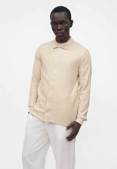 Homme portant une chemise beige texturée à manches longues boutonnée et un pantalon blanc, debout devant un fond blanc uni.