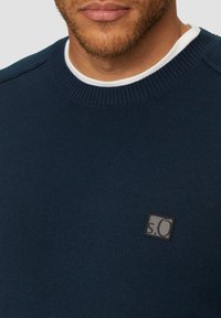 Navyblauwe gebreide trui met een geribde halslijn, voorzien van een klein vierkant logo-opstiksel in donkergrijs op de linkerborst.