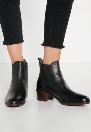 Bottines en cuir noir avec talons en bois bas portées avec un jean noir raccourci sur fond blanc.