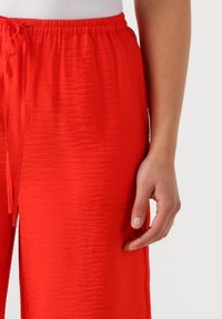 Rode, textuurachtige broek met elastische taille en trekkoorddetails. Een hand is naast het kledingstuk geplaatst, met een neutrale nagellak kleur.