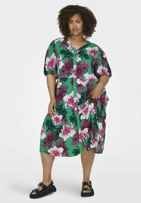 Blumen-Midi-Kleid aus grünem Stoff, mit großen pinken und weißen Blumen, Puffärmeln und einer Knopfleiste. Getragen mit schwarzen Sandalen.