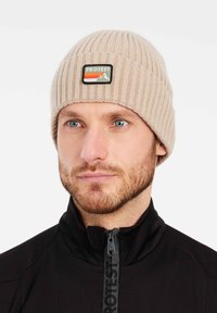 Beige knitted beanie with ribbed texture, featuring a rectangular black and green patch displaying mountains and the word "PROTEST."