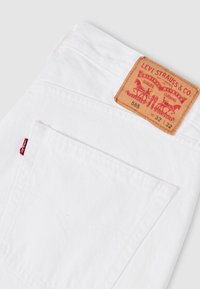 Λευκά τζιν Levi’s διπλωμένα, με εμφανή την πίσω τσέπη, την κόκκινη ετικέτα Levi’s και το ταμπελάκι από δέρμα σε ταμπά χρώμα με το λογότυπο της μάρκας και τις λεπτομέρειες μεγέθους.