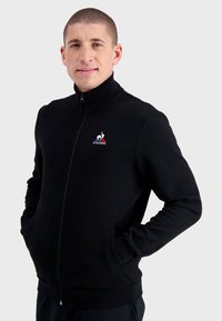 Giacca nera con zip realizzata in tessuto morbido, con colletto alto, tasche laterali e un piccolo logo ricamato sul petto.