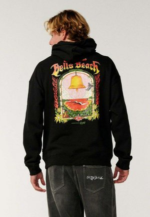 Joven con cabello rubio, vistiendo una sudadera negra con capucha con un gráfico colorido de Bell's Beach y jeans oscuros, de pie contra un fondo liso.