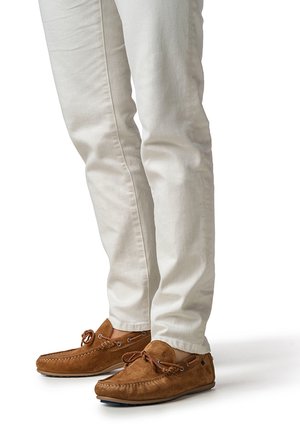 Man wearing light beige pants and brown suede moccasin loafers with laces, standing on a white background.