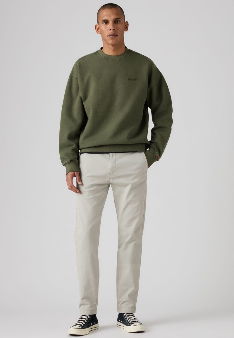 Homme debout portant un sweat-shirt vert olive, un pantalon beige clair et des baskets noires basses sur un fond gris uni.