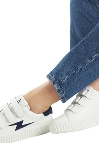 Baskets blanches avec trois bandes Velcro et accents navy, dotées d'une semelle en caoutchouc avec un motif texturé et un bout arrondi. Poignet en denim visible.