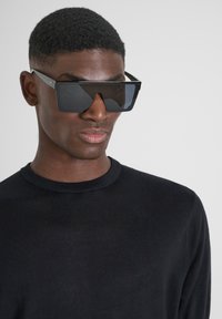 Occhiali da sole neri oversize con design angolare e lenti scure, abbinati a un maglione nero. Montatura elegante con accenti geometrici.