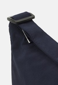 Borsa in tessuto blu navy con fibbia in plastica nera sulla tracolla, caratterizzata da una texture liscia e linee pulite in una forma triangolare.