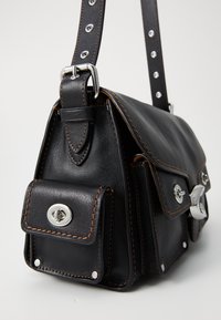 Sac à main en cuir noir avec quincaillerie en argent, présentant des coutures orange, une poche avant et une bandoulière réglable avec des clous décoratifs.