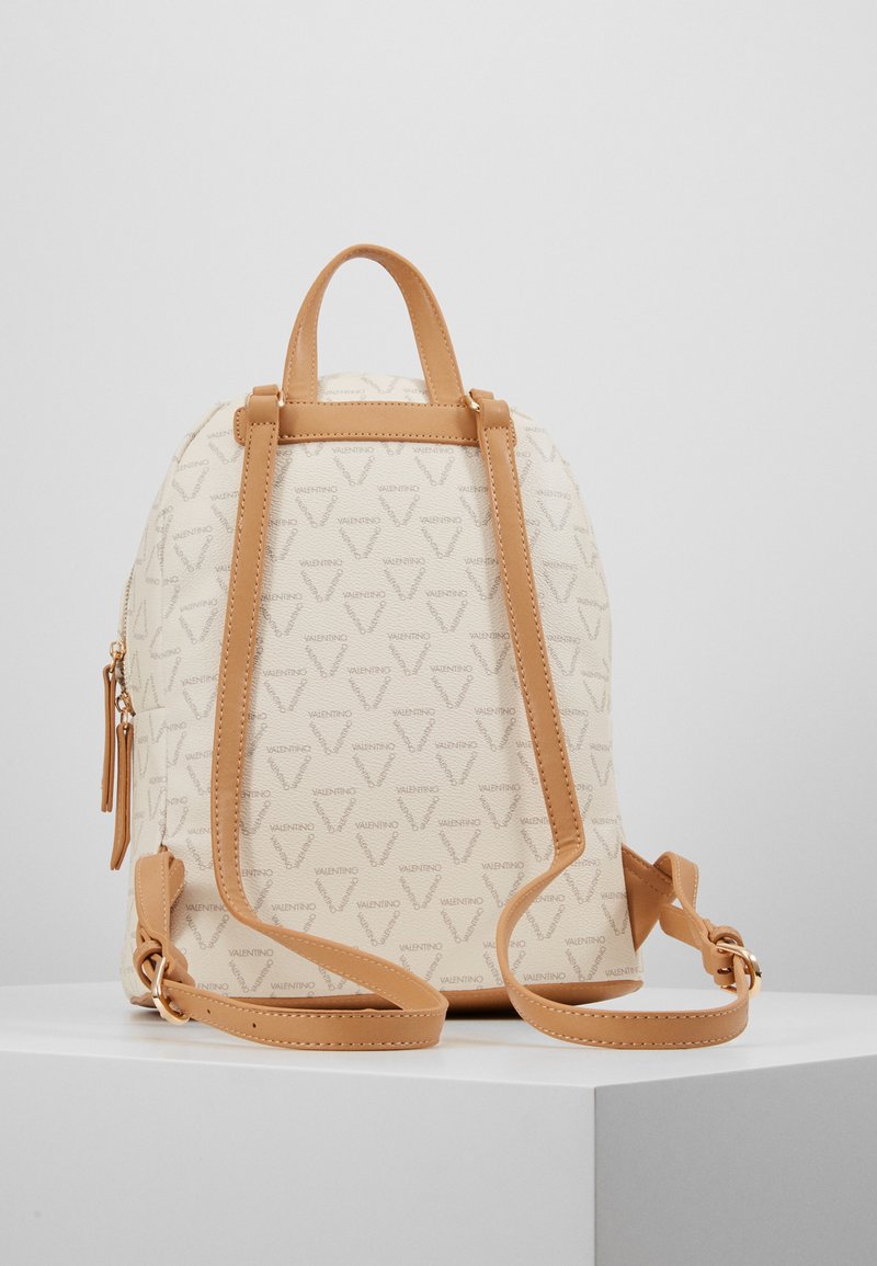 Mochila crema hecha de material texturizado, con un patrón de logo sutil. Acentos de cuero en color marrón y correas ajustables. Cierre de cremallera en el lateral.