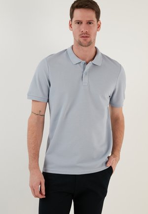 Lichtblauwe polo met korte mouwen, gemaakt van gestructureerde stof. Heeft een kraag en een knoopsluiting met twee knopen, gecombineerd met donkere broeken.