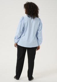 Blouse texturée bleu clair avec manches bouffantes, associée à un pantalon noir et des chaussures plates. La blouse a un ourlet arrondi et une empiècement au dos.