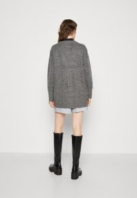 Grijze oversized cardigan met lange mouwen, gebreide textuur en zijsplitten, gecombineerd met een lichtblauw shirt en kniehoge zwarte laarzen.
