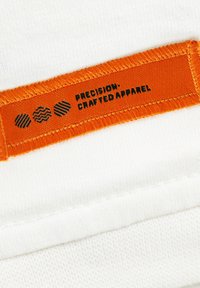 White fabric with a textured finish. Orange label features black text, "PRECISION: CRAFTED APPAREL," and three graphic shapes.